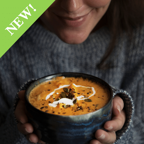 Roasted Butternut Squash Soup garnished with cream, pumpkin seeds and seasoning held by person in cozy blue sweater