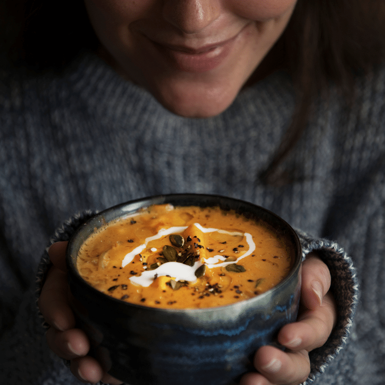 Roasted Butternut Squash Soup garnished with cream, pumpkin seeds and seasoning held by person in cozy blue sweater